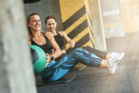 Two young happy athletic women in sportswear exercising with fitness ball at gym, working out togetherの写真素材
