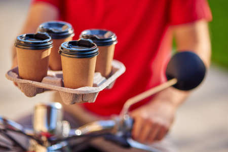 Male courier holding four coffee cups, delivering order by scooter to customerの写真素材