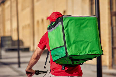 Rear view of a male courier in red uniform with thermobag riding bicycle along street on a sunny dayの写真素材