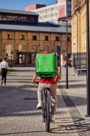 Rear view of a courier with thermo bag riding bicycle along tiled street on a sunny dayの写真素材