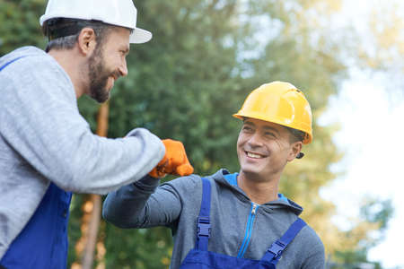 Team. Two positive young male engineers in hard hats smiling at each other, giving fist bump while working on cottage construction site outdoorsの写真素材