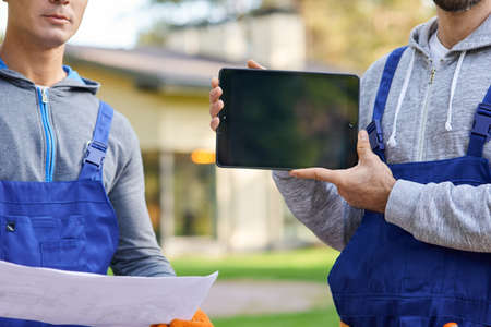 Cropped shot of young male builder showing screen of digital tablet pc with coworker holding...