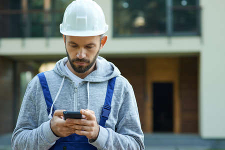 Young male builder in hardhat using smartphone, standing outdoors while working at cottage construction siteの写真素材
