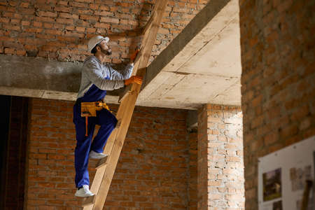Young male builder in blue overalls and hard hat looking focused, climbing up the ladder while...