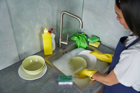 Make it clean. Young woman, cleaning lady in yellow gloves washing dishes with a sponge in the sink....