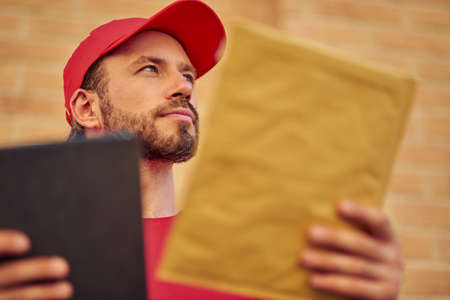 Close up shot of a young caucasian male courier holding small paper parcel and looking aside while standing outdoors, selective focusの写真素材