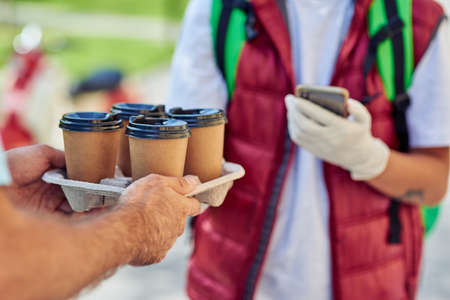 Male courier wearing protective gloves delivering four coffee cups to customer and using mobile app on his smartphone while standing on the sunny streetの写真素材