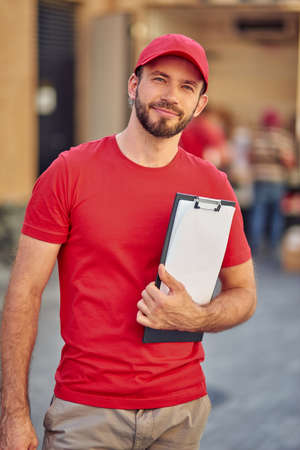 Young happy caucasian male courier in uniform holding folder with documents and smiling at camera...