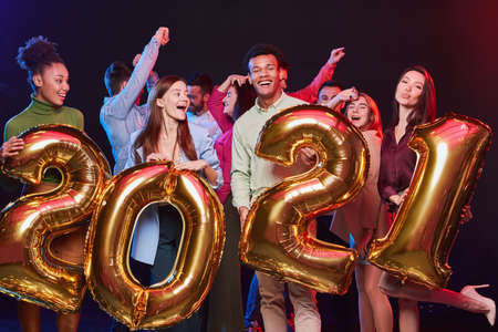 New Years Celebration. Group of young happy multiethnic people holding golden foil balloons in the form of numbers 2021 and having fun at Christmas partyの写真素材