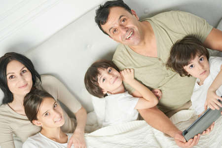 Together at home. Portrait of happy latin famile, little kids and parents smiling at camera while spending time together, staying in bed in the morningの写真素材