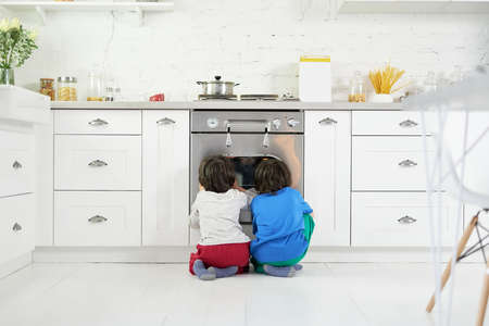 Cute little latin boys, twins watching pie baking in the oven, crouching down in the kitchen at homeの写真素材