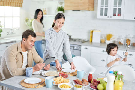 Middle aged latin man using tablet pc, waiting for dinner while his happy children and wife serving table in the kitchen at homeの写真素材