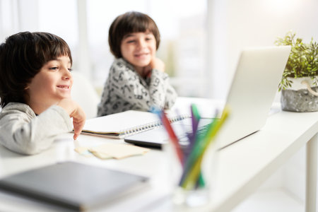 Two smiling little hispanic boys, twin brothers looking at the laptop screen during online lesson for children, sitting at the table at homeの写真素材