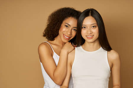 Portrait of two adorable mixed race young women with perfect skin smiling at camera while posing together, standing isolated over light brown backgroundの写真素材