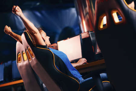 Side view of a young excited female cybersport gamer raising hands up, celebrating success while...