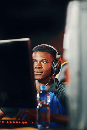 Fully concentrated young african guy, professional cybersport gamer wearing headphones sitting in gaming club or internet cafe and playing online video gamesの写真素材