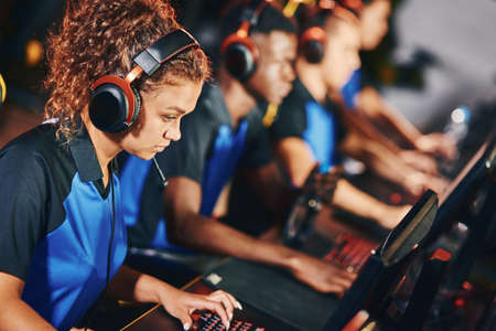 Focused female cybersport gamer wearing headphones participating in eSport tournament, sitting in a row with other team members and playing online video gamesの写真素材