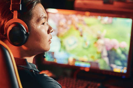 Close up shot of a focused asian guy, professional cybersport gamer wearing headphones playing...