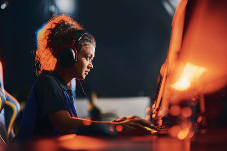 Side view of a young mixed race girl, female cybersport gamer wearing headphones playing online video games, participating in eSport tournamentの写真素材