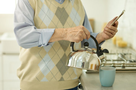 Aged man looking and smartphone and pouring water from kettleの写真素材
