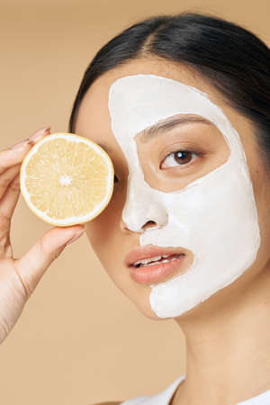 Beauty portrait of young woman with facial mask applied on half of her face holding a slice of lemon, posing isolated over beige backgroundの写真素材