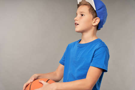 Cute boy basketball player standing against gray background