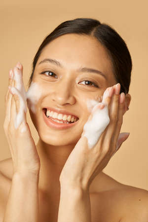 Beauty portrait of joyful young woman smiling at camera while applying gentle foam facial cleanser...