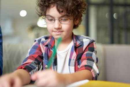 Portrait of concentrated latin boy in glasses writing while sitting at the desk and doing homework at homeの写真素材