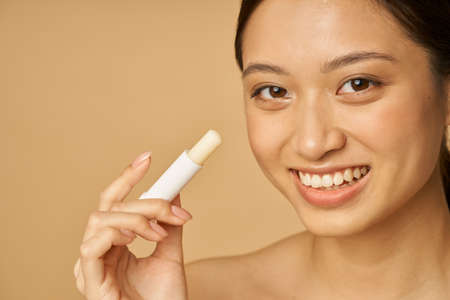 Face closeup of adorable young woman with beautiful smile looking at camera and holding lip balm, posing isolated over beige backgroundの写真素材