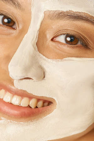 Happy young woman with facial mask applied on half of her face receiving spa treatments, smiling at...