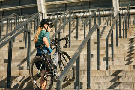 Sportive young woman, professional female cyclist looking away while coming up the steps with bicycle, training outdoorsの写真素材
