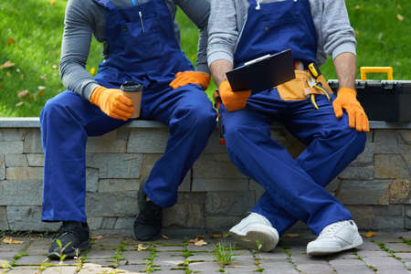 Cropped shot of two young workmen in uniform taking a break, sitting outdoors having coffee and holding papers while working on construction projectの写真素材