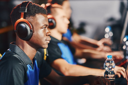 Focused african male cybersport gamer wearing headphones participating in eSport tournament, sitting in a row with other team members and playing online video gamesの写真素材