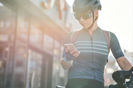 Female cyclist in protective gear using smartphone while riding bicycle in city center. Sportswoman training, exercising outdoorsの写真素材
