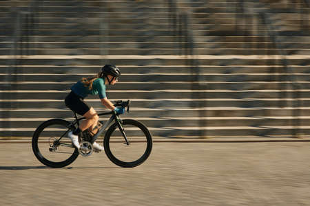 Side view of professional female cyclist in cycling garment and protective gear riding bicycle in city, rushing and passing buildings while training outdoors on a daytimeの写真素材