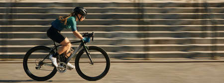 Website header of Side view of professional female cyclist in cycling garment and protective gear riding bicycle in city, rushing and passing buildings while training outdoors on a daytimeの写真素材