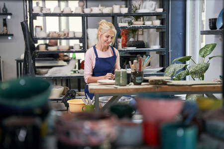 Handcraft. Relaxed mature woman in blue apron looking focused while creating handmade clay ceramics in pottery workshop studioの写真素材