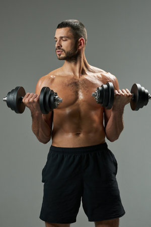 Focused adorable Caucasian man in sportswear lifting weights isolated on grey backgroundの写真素材