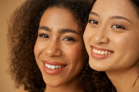 Face closeup of two joyful attractive mixed race young women with perfect smile posing for camera isolated over beige backgroundの写真素材