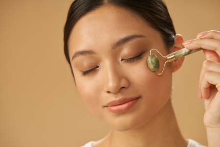 Portrait of relaxed young woman using jade roller for massaging her face, posing with eyes closed isolated over beige backgroundの写真素材