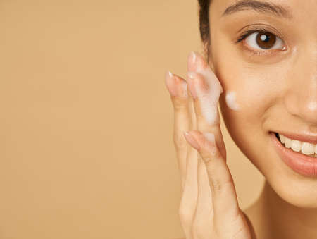 Studio half face portrait of beautiful young woman smiling at camera while applying gentle foam facial cleanser isolated over beige backgroundの写真素材