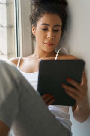 Relaxed young woman reading a book using tablet pc while sitting by the window at homeの写真素材