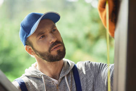 Close up portrait of focused young builder wearing blue cap and overalls using measuring tape while working on cottage construction on a sunny dayの写真素材