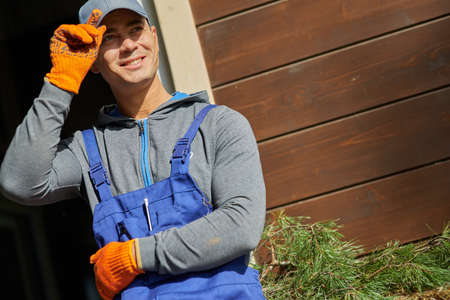 Portrait of joyful young male builder wearing blue overalls and cap smiling while taking a break,...