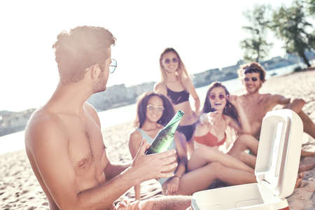 Enjoying carefree time with friends. Cheerful young people spending nice time together while sitting on the beach and drinking beerの写真素材