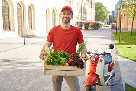 Happy courier in uniform holding grocery box with fresh vegetablの写真素材