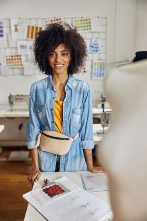 Professional African-American seamstress in denim shirt with beige belt bag stands at table in...