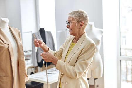Joyful woman designer with tape measure stands near mannequin with beige jacketの写真素材