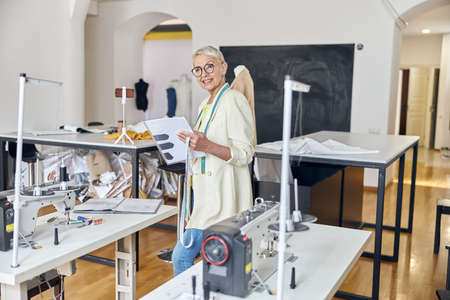 Positive mature tailor with stylish glasses holds sketchbook standing in modern workshop