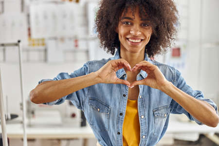 Smiling African-American fashion designer in denim shirt shows heart in sewing workshopの写真素材